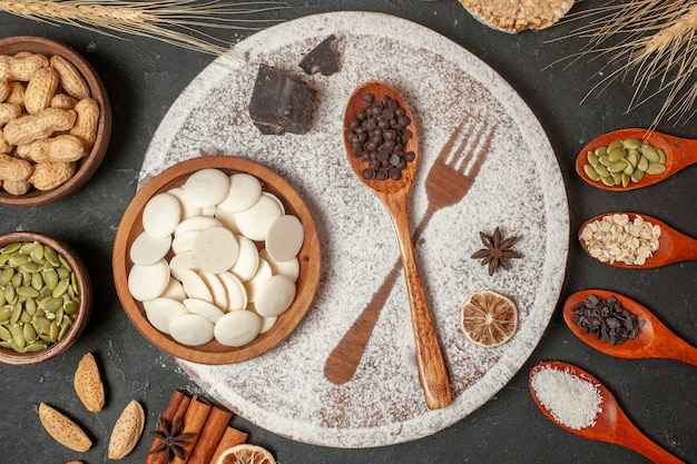 bovenaanzicht witte chocolade rondjes in kom vork opdruk met poedersuiker houten lepel op bord noten in kommen kaneelstokjes houten lepels op tafel