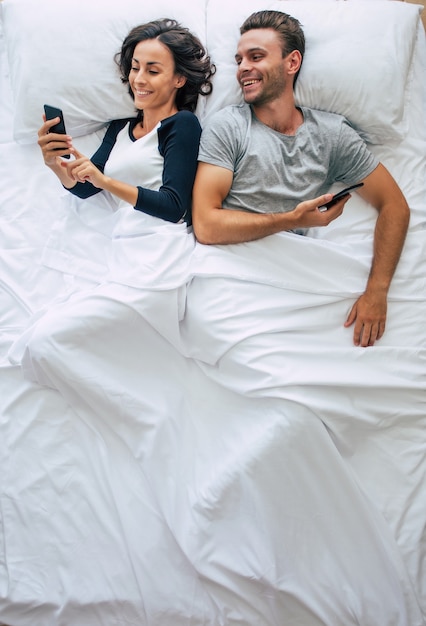 Bovenaanzichtfoto van de mooie vrouw en de knappe jongeman gebruiken smartphones terwijl ze in een groot wit bed liggen. online chatten. de foto hierboven van het paar.