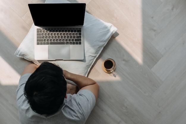 Foto bovenbeeld van een aziatische man die met een computer op de vloer ligt terwijl hij aan een laptop werkt met koffie en typen