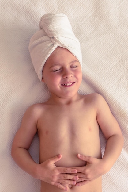 Bovenbeeld van een verheugd klein kind met naakte romp en een handdoek op het hoofd dat met gesloten ogen op een witte deken ligt