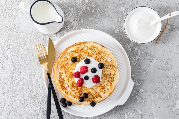 Bovenbeeld van vers gebakken pannenkoeken met zure room en bessen op een wit bord met bestek Een traditioneel ontbijt voor een vakantie in Rusland