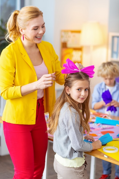 髪に頭を下げます。彼女のかわいい女子高生の髪にピンクの紙の弓を置くブロンドの髪の先生の笑顔