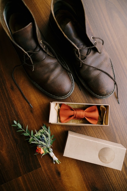 Bow tie in a box for a groom with a boutonniere and mens boots with untied laces on a brown floor