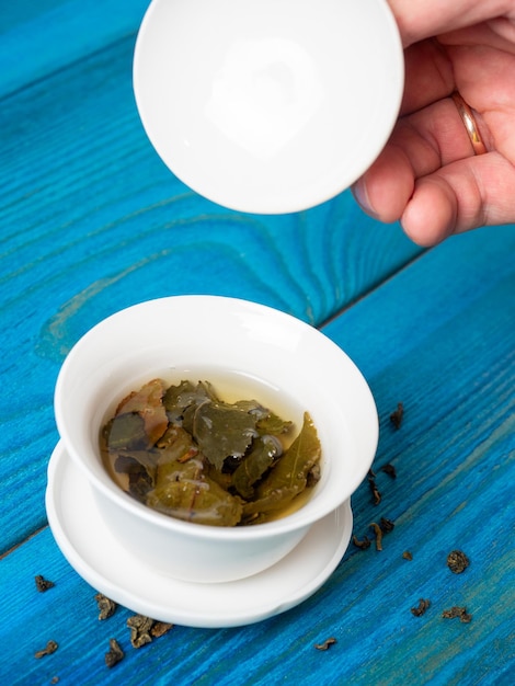 Una ciotola di tè preparato su uno sfondo di legno blu. una mano solleva il coperchio della ciotola. le foglie aperte di tè a foglia larga. tè cinese. tea party. foto verticale