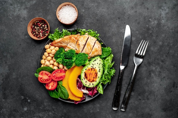 Foto bowl buddha. pollo, broccoli, ceci, zucca, avocado, carota, pomodoro, lattuga su un piatto con coltello e forchetta