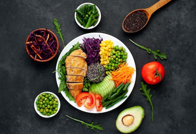 bowl buddha.quinoa, chicken breast, arugula, avocado, red cabbage, carrot, green peas, corn, tomato, green beans in a white plate.