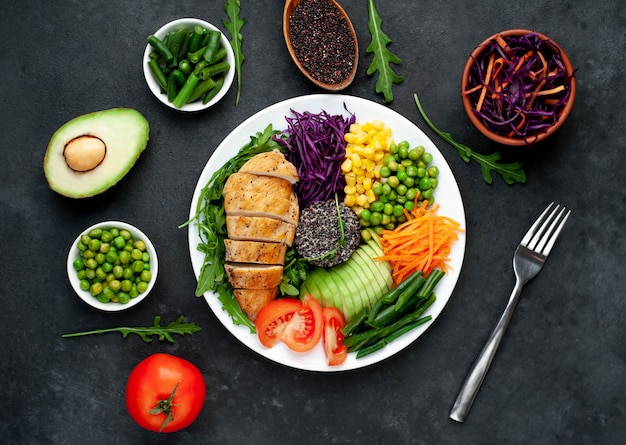 bowl buddha.quinoa, chicken breast, arugula, avocado, red cabbage, carrot, green peas, corn, tomato, green beans in a white plate.