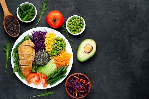 bowl buddha.quinoa, chicken breast, arugula, avocado, red cabbage, carrot, green peas, corn, tomato, green beans in a white plate.