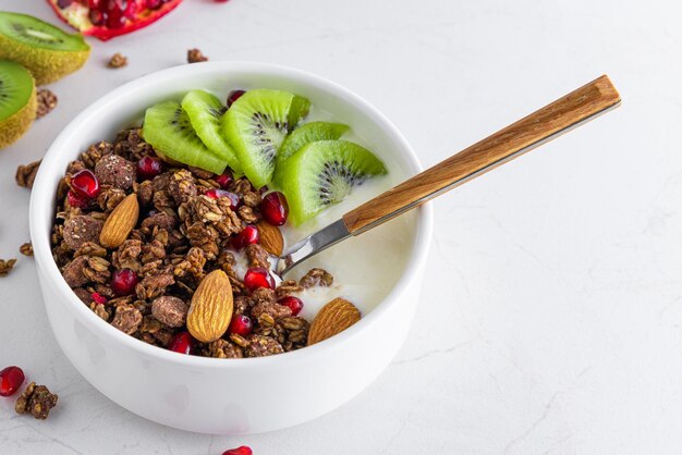 Bowl of chocolate oat granola with yogurt fresh kiwi fruit pomegranate seeds nuts and spoon on white marble table