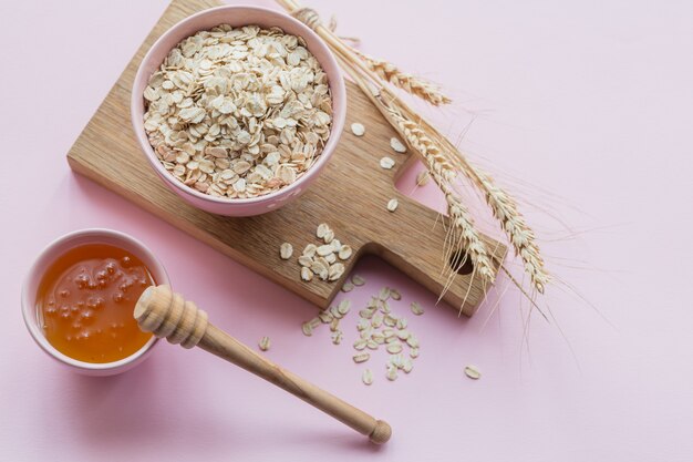 Bowl of dry oat flakes with honey and ears of wheat on light pink background. Cooking oats porridge concept