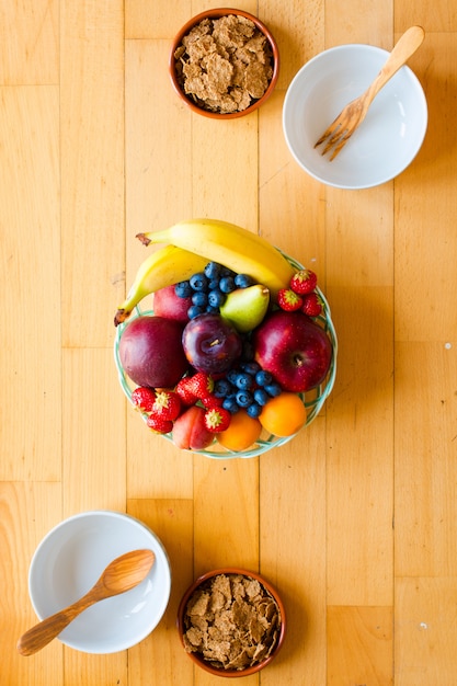 Bowl of fresh fruit with banana, apple, strawberries, apricots, blueberries, plums, whole grains, forks, top view