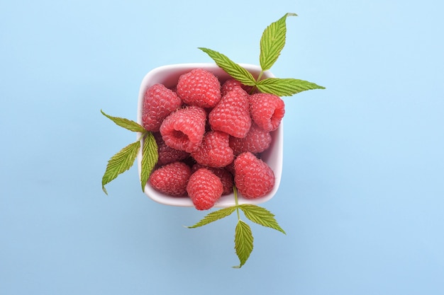 Bowl of fresh raspberries with green leaves on blue background. Top view.