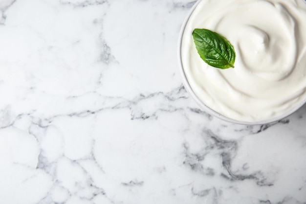 Bowl of fresh sour cream with basil on white marble table top view Space for text