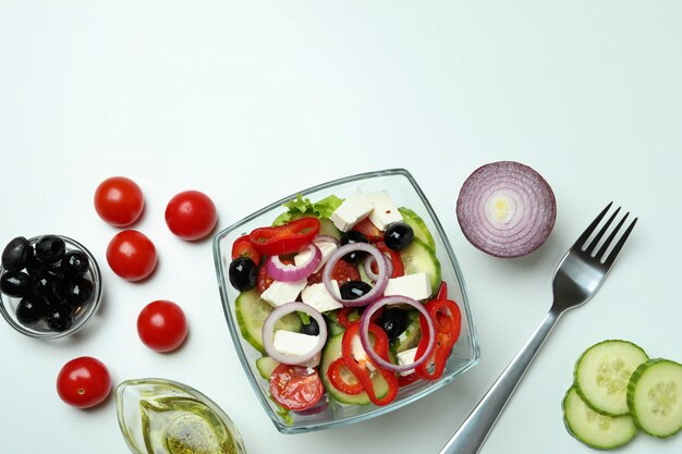 Bowl of greek salad and ingredients on white surface