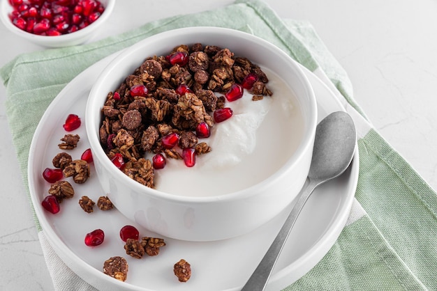 Bowl of greek yogurt with chocolate oat granola and pomegranate seeds on white marble table