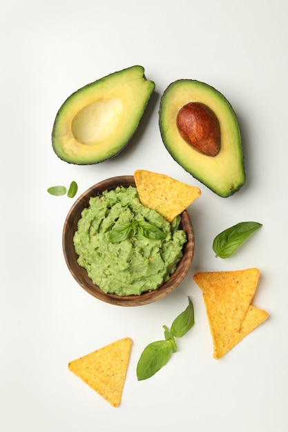 Bowl of guacamole, avocado, chips and basil on white background, top view