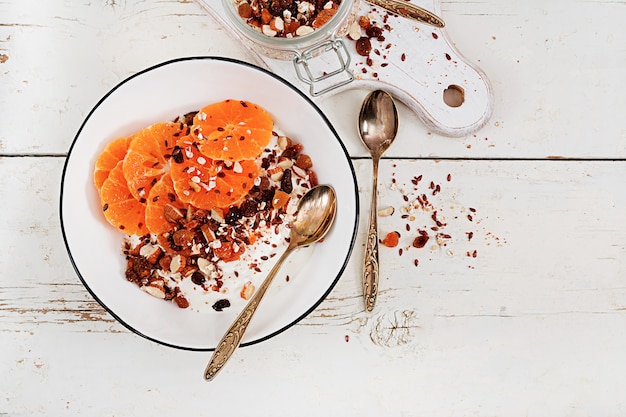 Bowl of homemade granola with yogurt and tangerine on white wooden table.