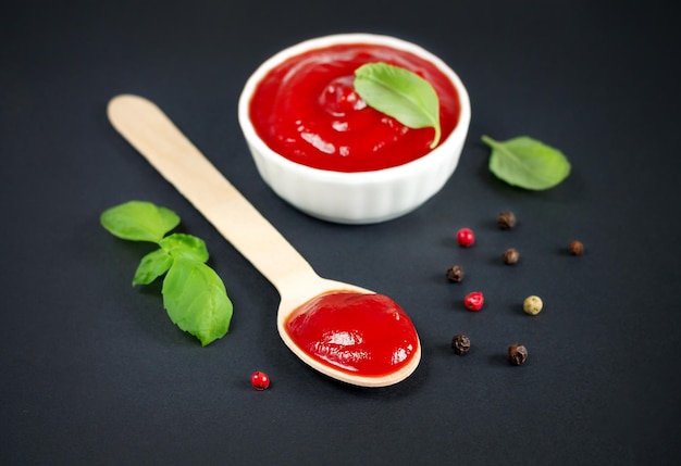 Bowl of ketchup and wooden spoon of red tomato sauce next to basil and spices on black kitchen table