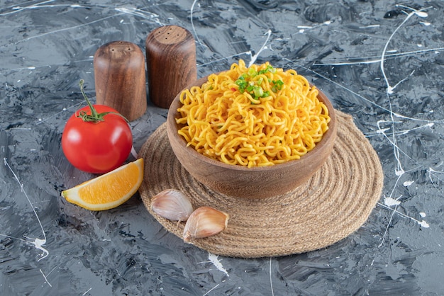 A bowl of noodle on a trivet next to tomatoes lemon and garlic on the marble background