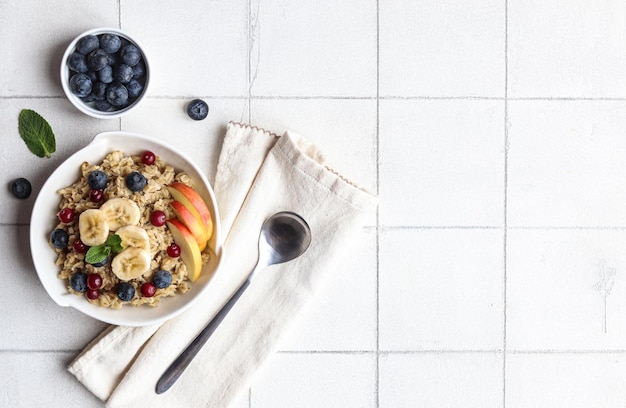 A bowl of oatmeal with fruit and a spoon on a white tile background.