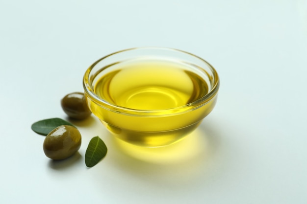 Bowl of oil, olives and leaves on white surface