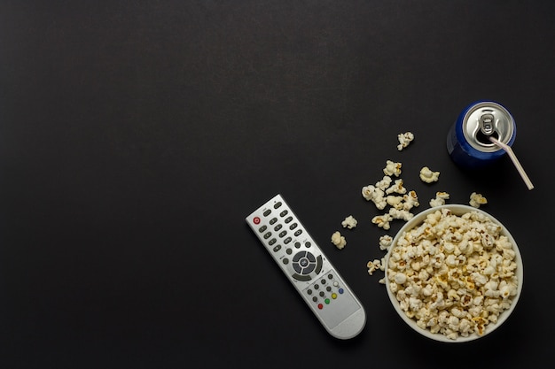 A bowl of popcorn, a tv remote, a can of drink on a black background. the concept of watching tv, film, tv series, sports, shows. flat lay, top view.