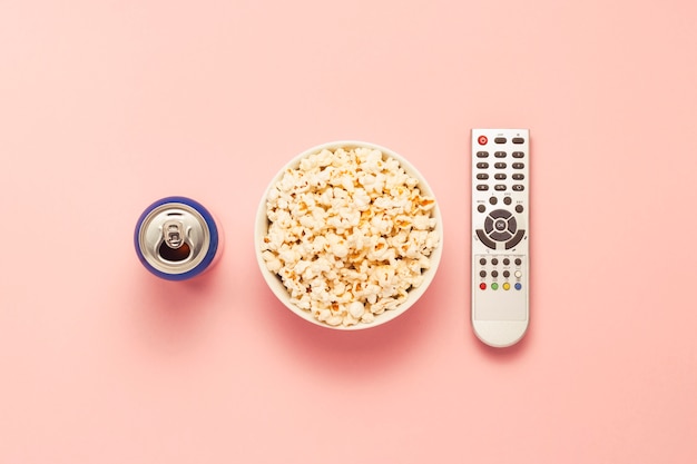 A bowl of popcorn, a TV remote, a jar of drink on a pink background. The concept of watching TV, film, TV series, sports, shows. Flat lay, top view.