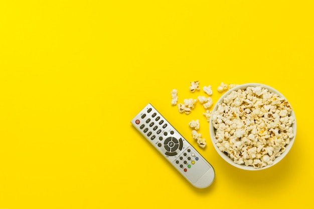 A bowl of popcorn and TV remote on a yellow background. The concept of watching TV, film, TV series, sports, shows. Flat lay, top view.