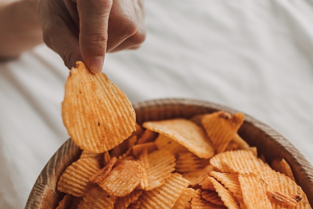 Bowl of potato chips on white bed with hand picking up