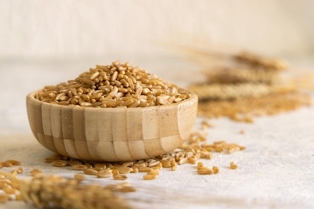 Bowl of raw dry rye grain on white table against rye ears close up