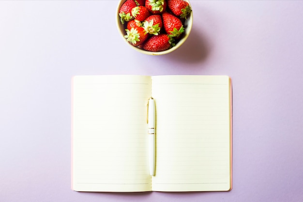 A bowl of ripe red strawberries stands next to an open notepad 