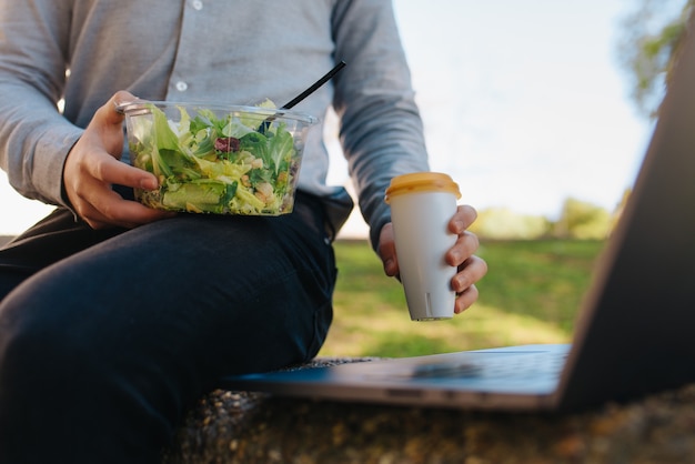 Photo a bowl of salad on the legs of an unrecognizable man well dressed grabbing a cup of takeaway coffee using his laptop on a cobblestone floor. close up