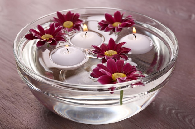 Bowl of spa water with flowers and candles on wooden table closeup