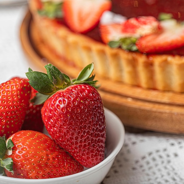 A bowl of strawberries next to a pie with a cake in the background.