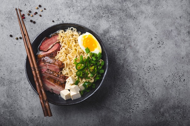 Bowl of tasty Asian noodle soup ramen with broth, tofu, pork, egg on grey rustic concrete background, space for text, close up, top view. Hot tasty Japanese ramen soup for dinner with copy space