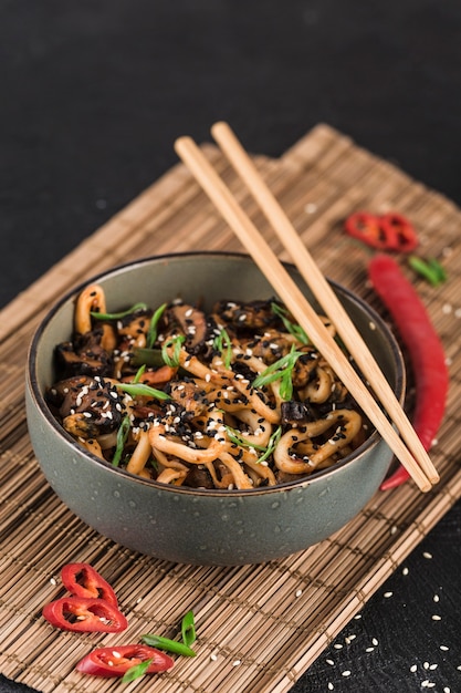 A bowl of udon noodles with squid rings and mushrooms with chopsticks and chili pepper on a dark surface