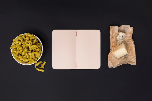 Bowl of uncooked pasta; blank open diary and cheese on kitchen top