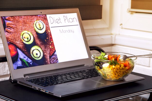 Bowl of vegetable salad near the laptop on the desktop. On the screen - planning a diet for weight loss from Monday