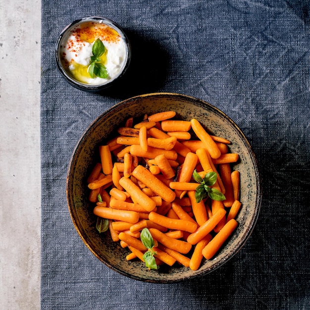 Bowl of whole baked baby carrot with yogurt sauce on blue tablecloth over grey background. Vegan dish. Flat lay, copy space