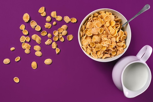 Bowl with corn flakes jug of milk and spoon on purple background top view