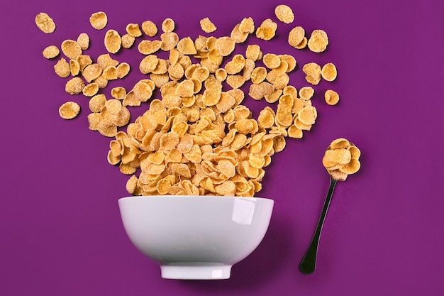 Bowl with corn flakes and spoon on purple background top view