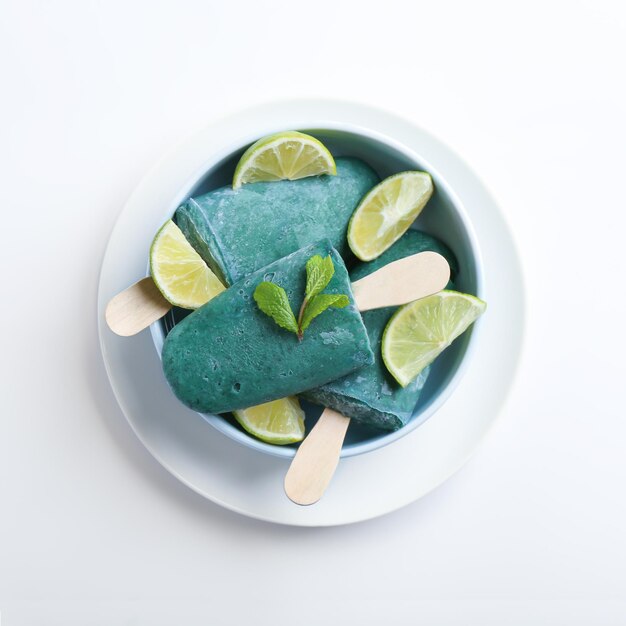 Bowl with delicious spirulina popsicles and lime slices on white background top view