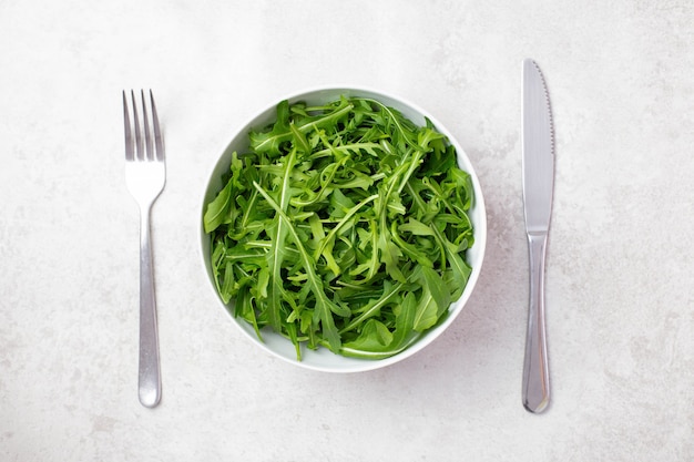 Bowl with fresh green and healthy arugula rucola salad with cutlery, fork and knife on light gray surface