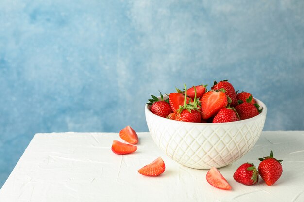 Photo bowl with fresh strawberries on white table against blue background, space for text. summer sweet fruits and berries