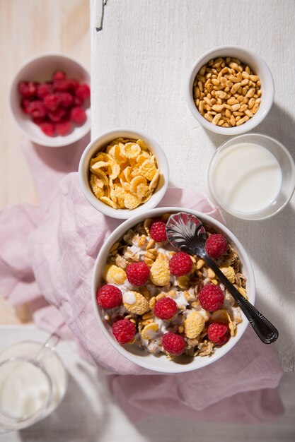 Bowl with oatmeal, corn flakes, raspberry and a glass of milk on a white wooden