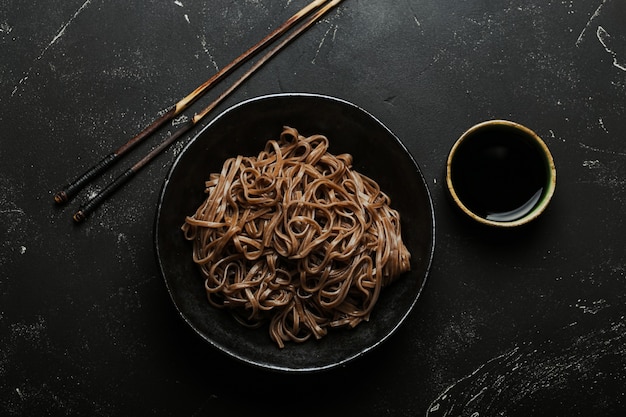 Bowl with plain soba noodles on dark black stone background from above, noodles for cooking Chinese Thai or Japanese dish with soy sauce