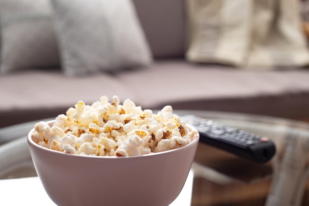 Bowl with popcorn and tv remote control on glass table near sofa