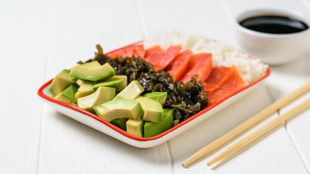 Bowl with rice, avocado, salmon and kelp on a white table.