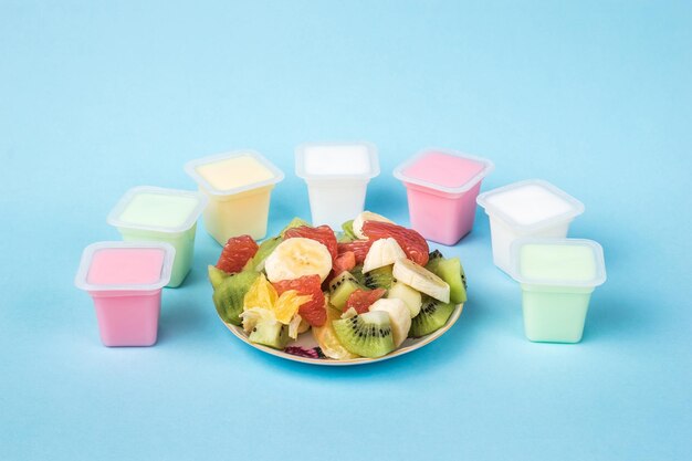 A bowl with sliced fruit and cups with multicolored yogurt on a blue background. Milk and fruit food.