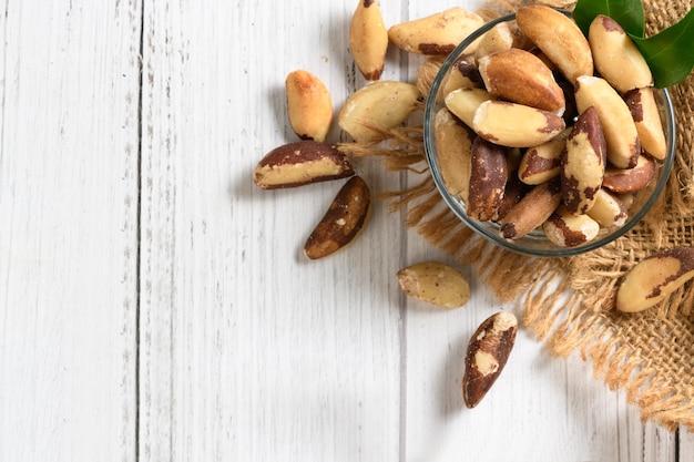 Bowl with tasty Brazil nuts on sack and space for text on white wooden background